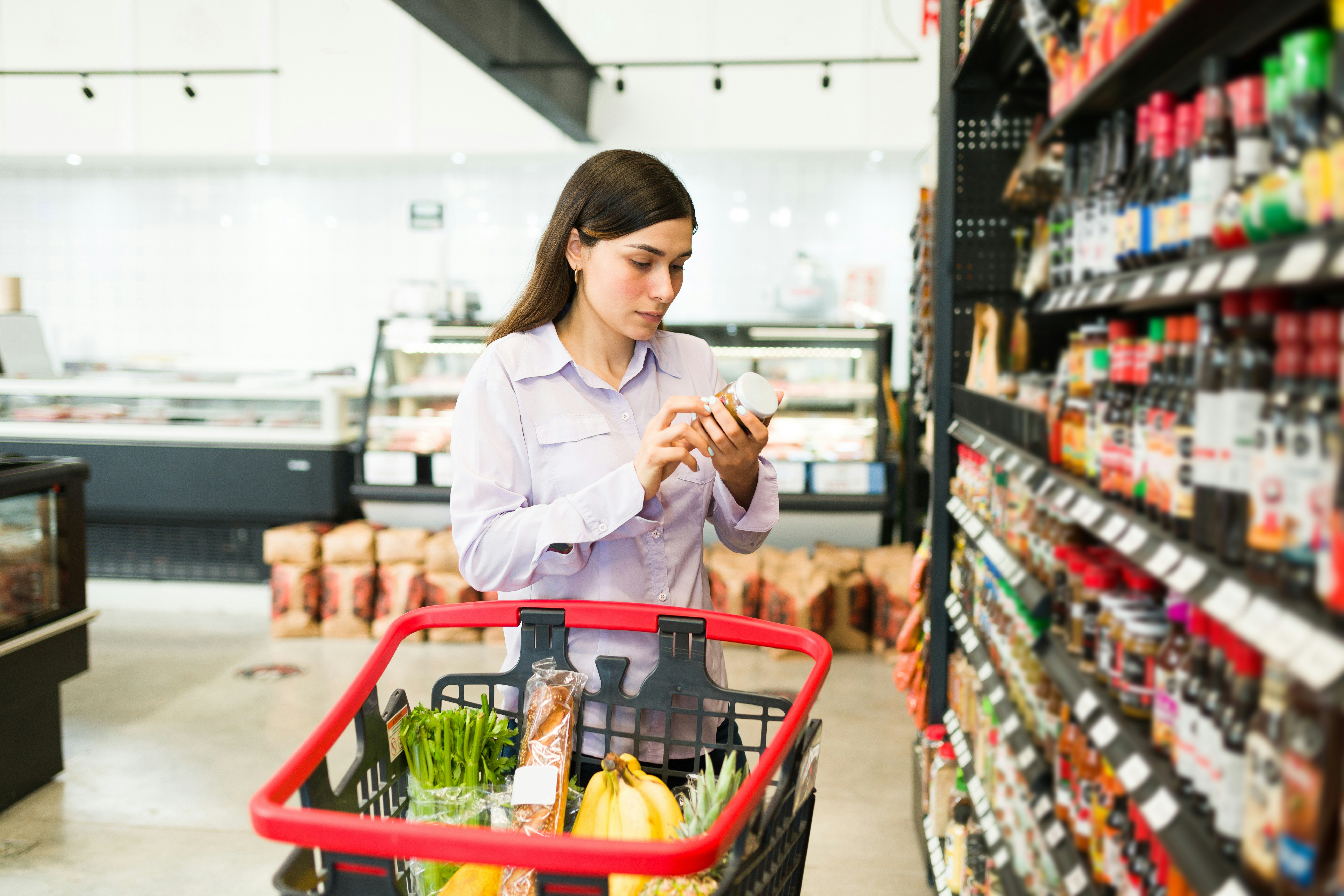 Woman shopping in a supermarket, checking a product label while standing beside a grocery cart filled with fresh produce and packaged food.