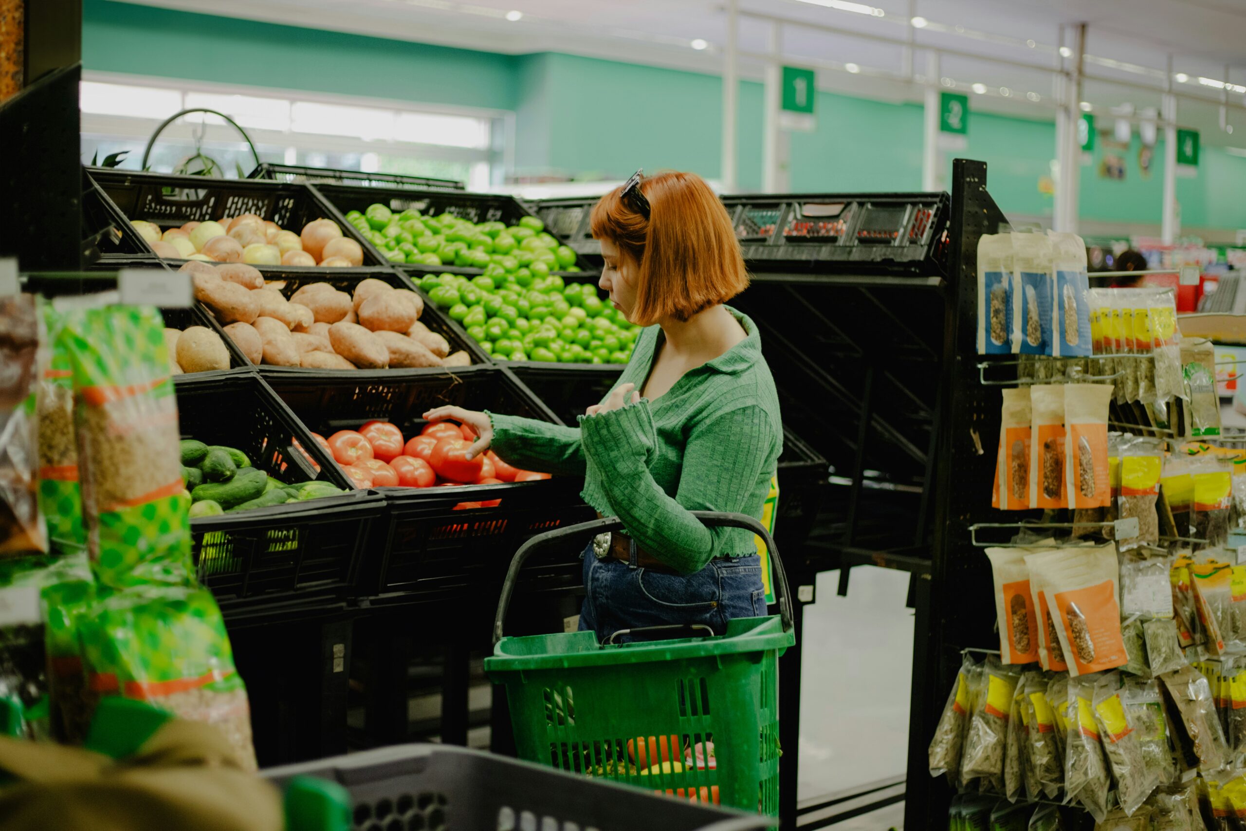 Woman shopping for fresh vegetables in a supermarket, selecting tomatoes from a produce display with a green shopping basket, illustrating grocery retail and everyday purchasing decisions.